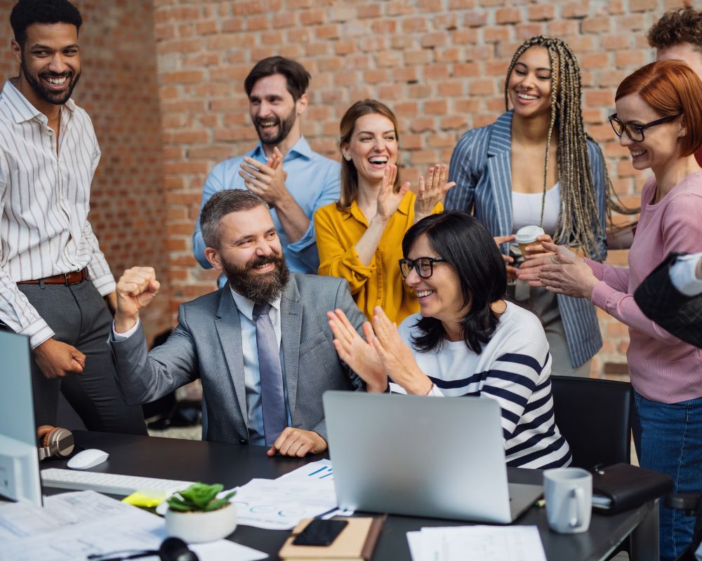 Group of diverse colleagues celebrating a success in a modern office, with one man in a suit raising his fist in excitement while others clap and smile around him. | Done-for-You Content Writing Services by Your Perfect Solutions